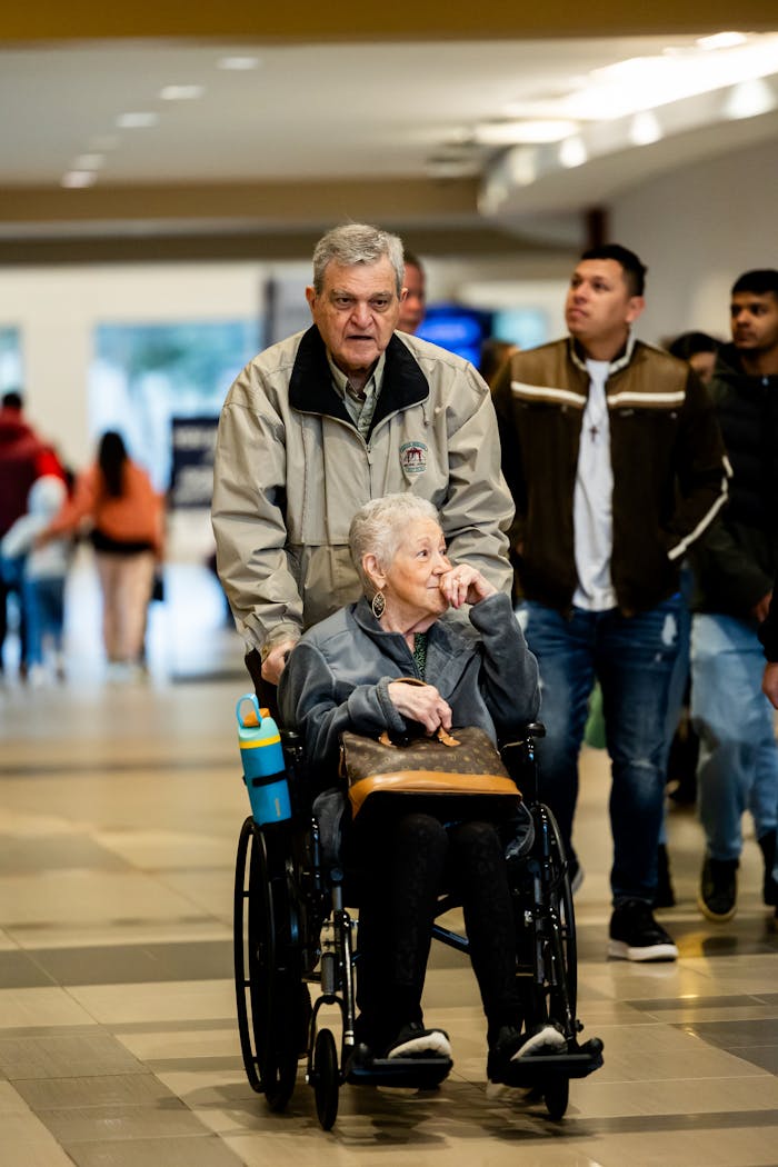 Senior woman in a wheelchair being assisted by a man in a bustling shopping mall.